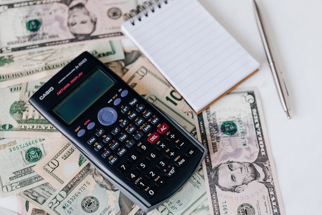 A calculator, a notepad, and a pen on a pile of dollar bills.