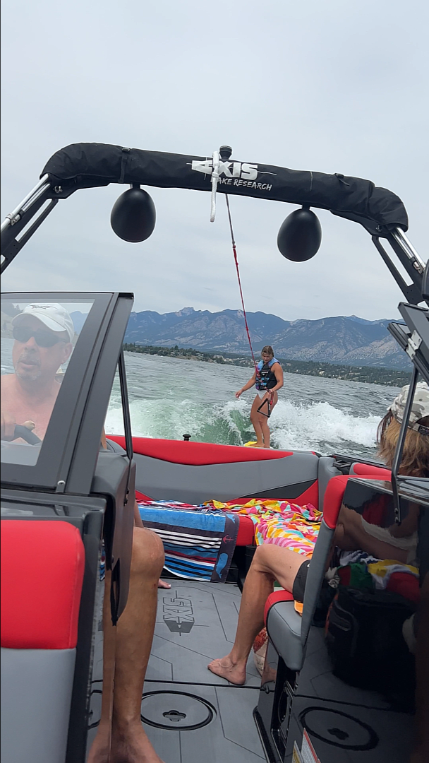Summer fun at Lake Windermere Lakefront with a view from a boat showing a person wake surfing on the water, with the rugged mountains in the distance under a partly cloudy sky, reflecting the active lakefront lifestyle