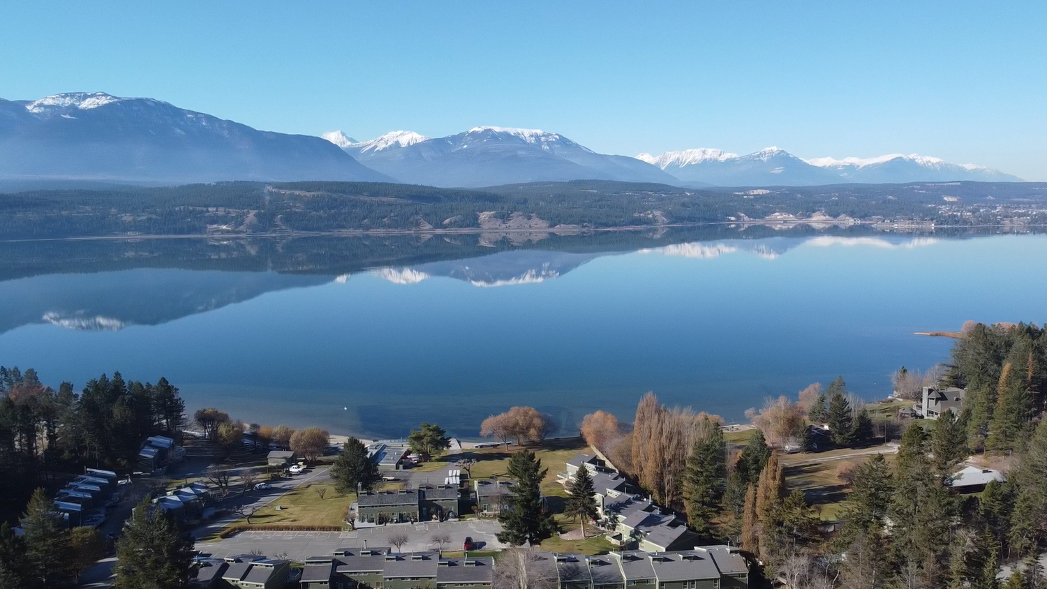 Aerial view of Lake Windermere's serene lakefront, showcasing the mirror-like reflection of surrounding mountains on the calm water, with lush trees and a community of houses along the shoreline.