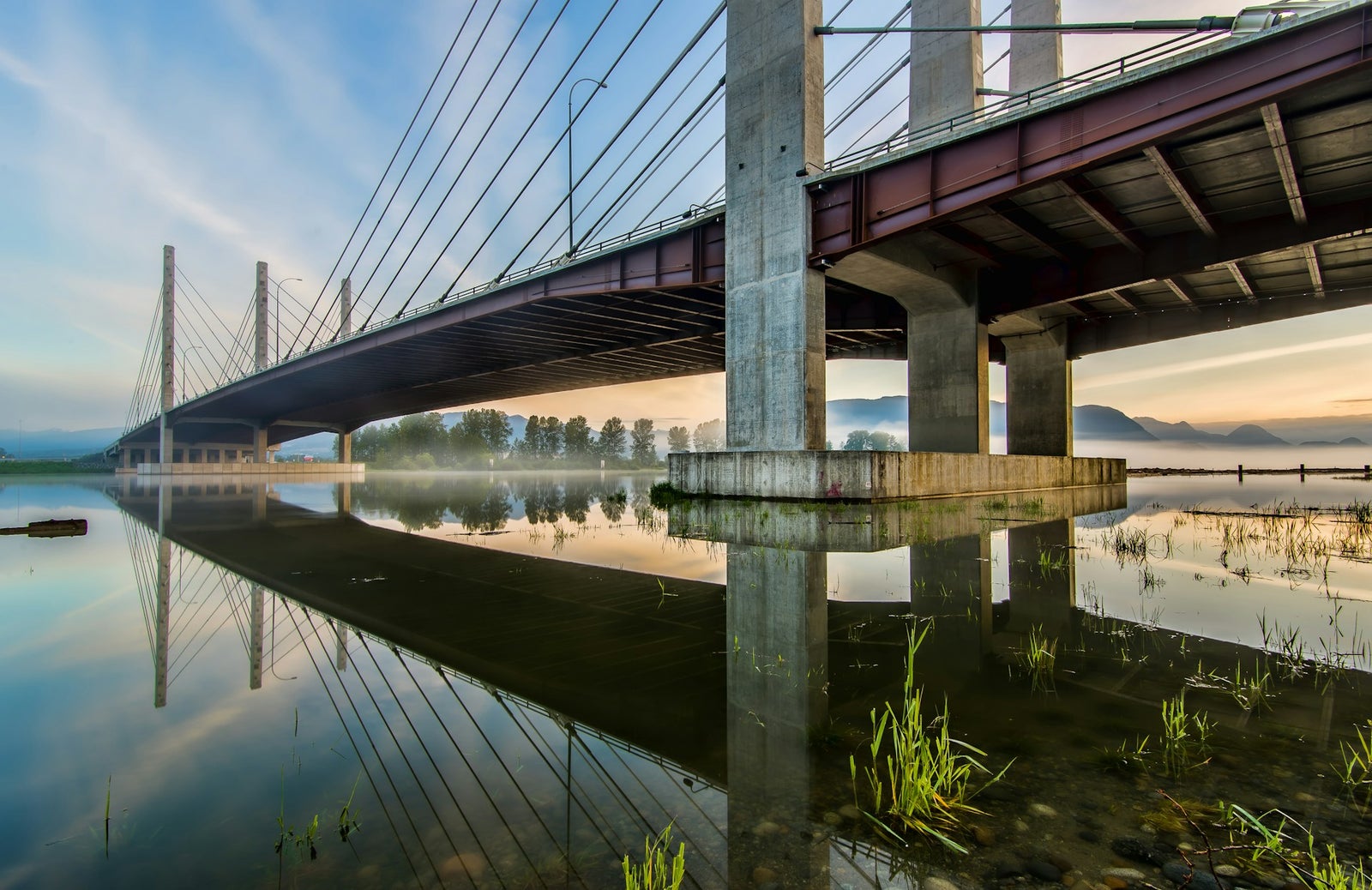 Pitt River Bridge at sunrise in Port Coquitlam, BC, reflecting on the Fraser River with mountains in the background.